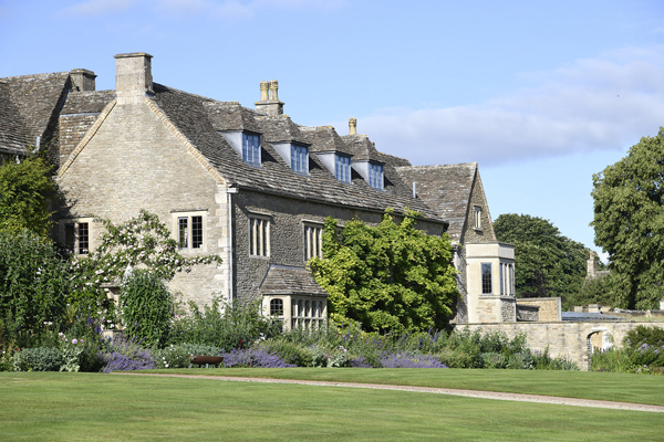 Sunlit Cotswold stone exterior of Whatley Manor with herbaceous flowerbeds and mullioned windows